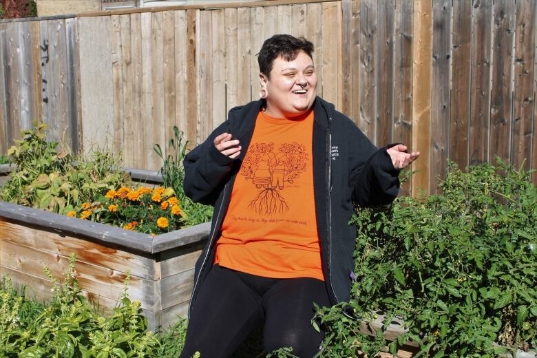 A person with short hair and wearing an orange shirt smiles as they speak while sitting in a garden with several raised plant beds.