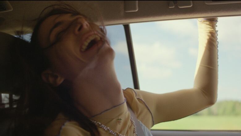 A woman rides as a passenger in a car laughs as the breeze through the open window blows her hair.