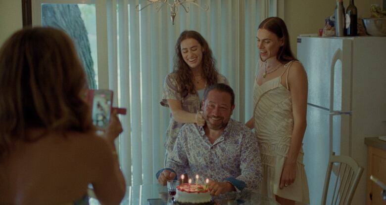 Two women stand smiling behind a man seated in front of a cake with candles.