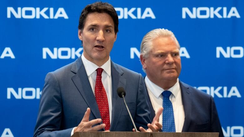 A politician speaks at a lectern in front of a blue corporate backdrop while another listens behind him.