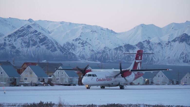 An airplane taxis on a snow-covered runway with large, ridig, snow-capped mountains in the background.