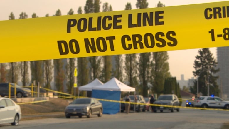 Yellow police tape reading 'Police line do not cross' is in the foreground with a hazy sky, tall trees and a police tent in the background.