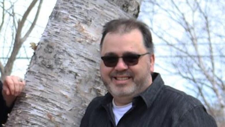 A man wearing dark glasses stands in front of a tree.