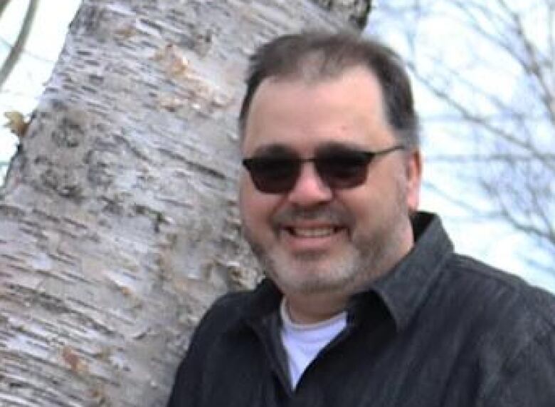 A man wearing dark glasses stands in front of a tree.