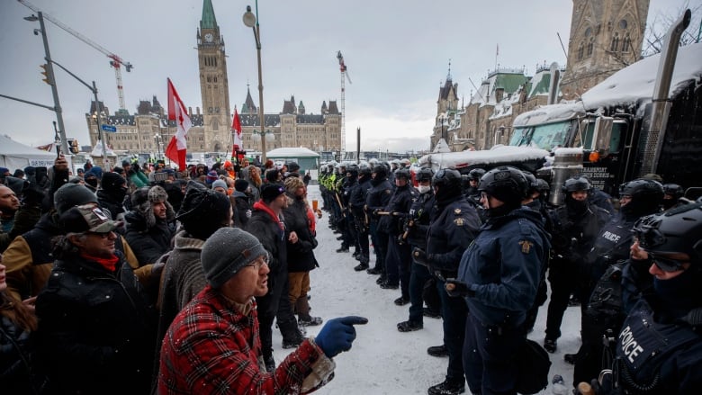 A row of protesters yells at a row of police officers in front of a legislature in winter.