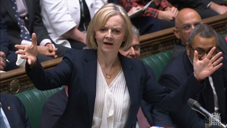 A woman with shoulder-length blond hair wearing a blue blazer speaks with her arms raised in the British Parliament. 