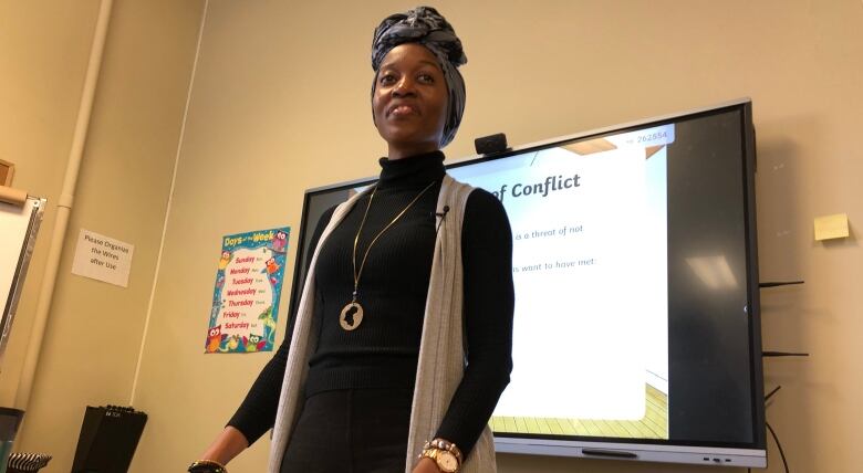 A smiling woman stands at a desk, with a large screen showing a presentation behind her.