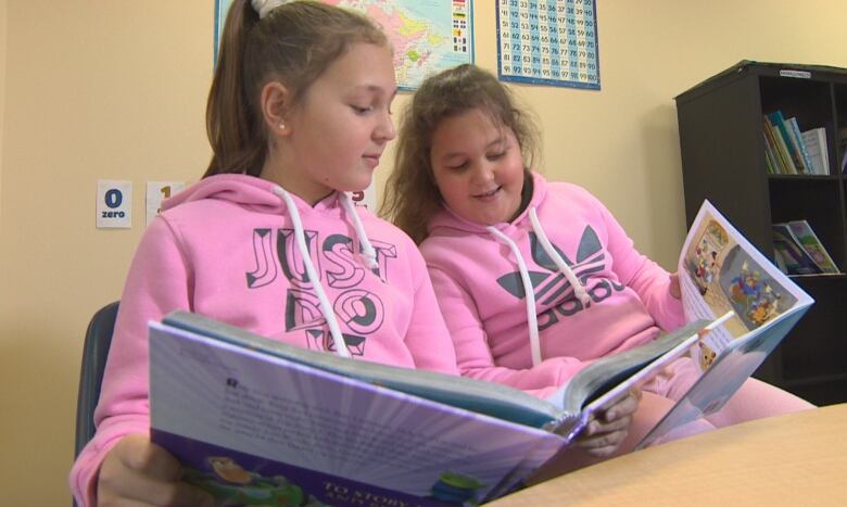 Two young girls in pink sweatshirts sit beside each other at a table, reading picture books.