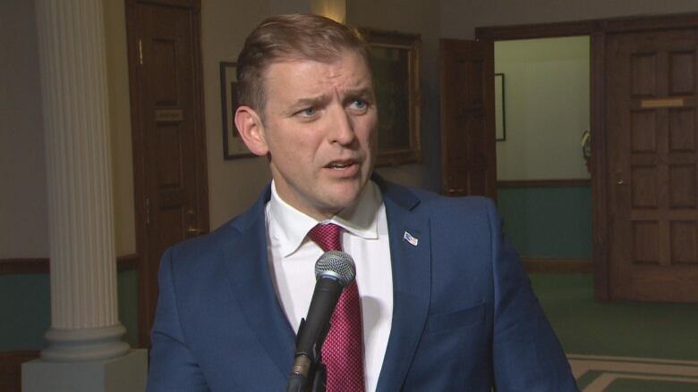 A man in a navy blue suit and maroon tie stands in front of a microphone. 