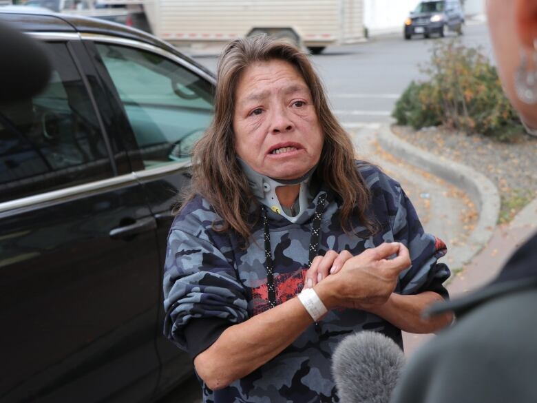 A woman with shoulder-length brown hair wears a neck brace and a hospital bracelet as she speaks to a reporter outside a hospital building. 