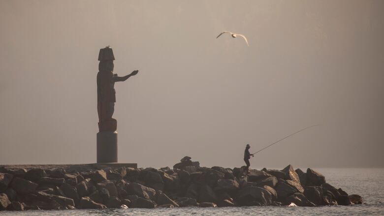 A lone figure is seen fishing at the end of a rock pier and their figure is blurred by hazy skies.