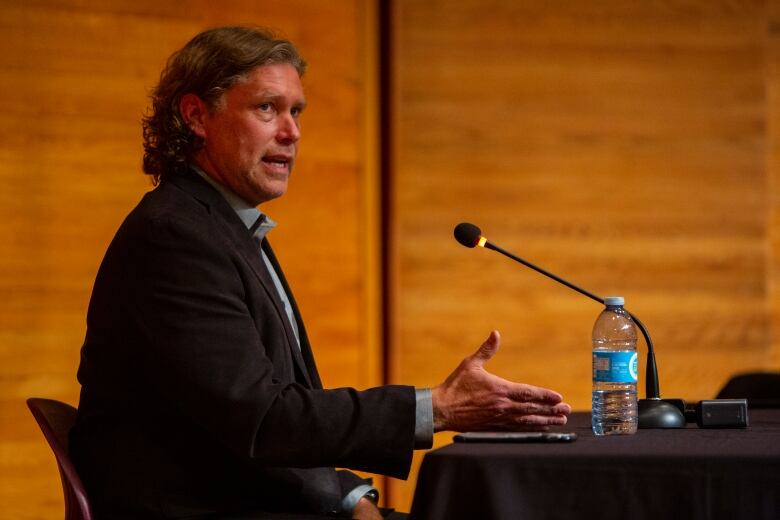 A man with curly hair sits talking at a table.