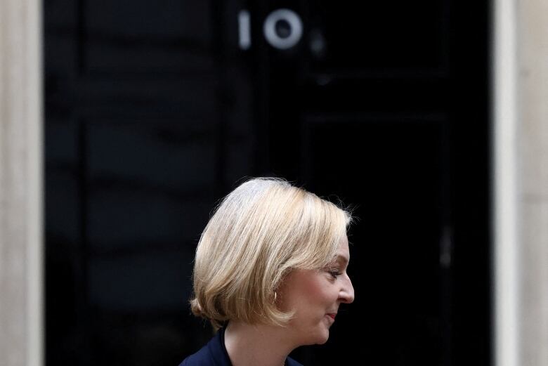 A middle-aged woman with a short blonde bob pictured in profile from the neck up. The black door of No. 10 downing street looms behind her, and she takes up a small portion of the bottom of the frame. 