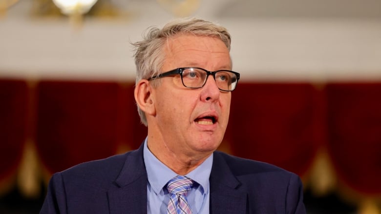 A man in a blue suit, with a blue shirt and plaid tie with glasses speaks.