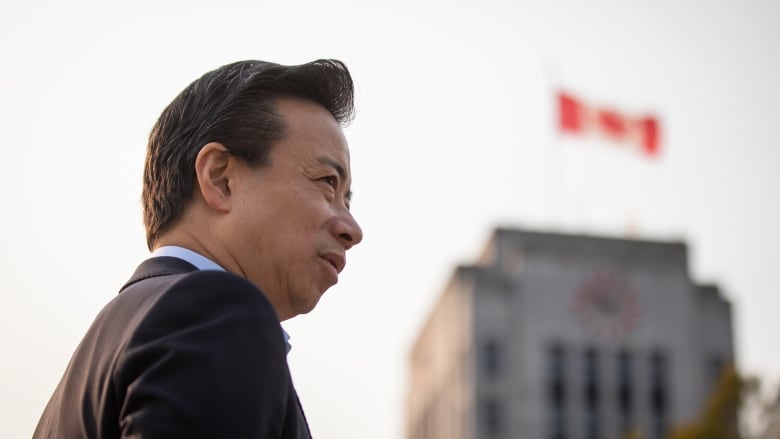 A man stands in profile with Vancouver city hall and a Canadian flag in soft focus to the right.