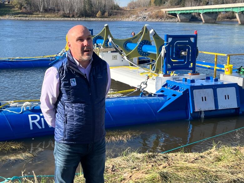 A man stands on the shore of the Winnipeg River, in front of a large piece of equipment floating on pontoons.