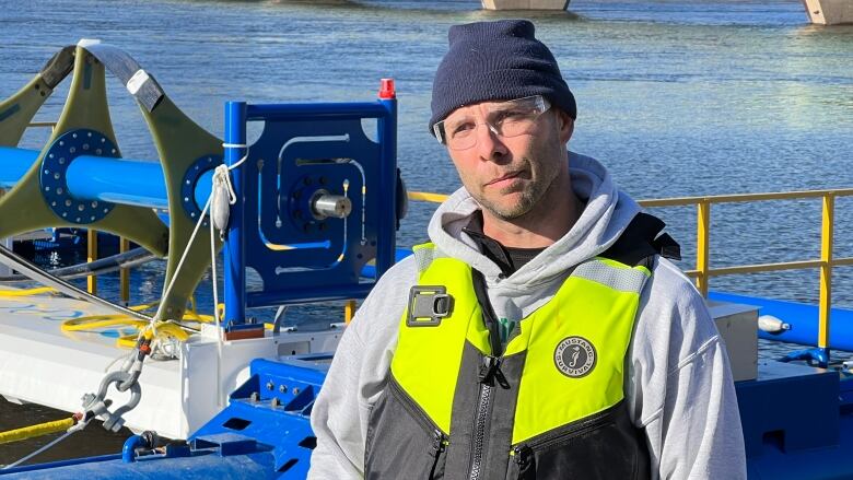 A man in a lifejacket and safety glasses stands on the shore of the Winnipeg River, in front of a large piece of equipment floating on pontoons.