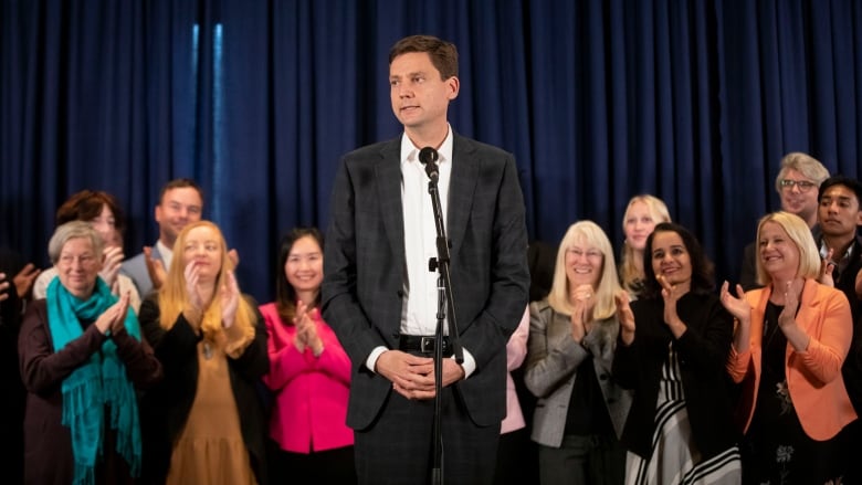 A white man speaks in front of clapping supporters.