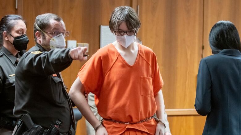 A teenage boy wearing glasses, a white mask over his face and dressed in an orange prison jumpsuit is led out of a court with his hands cuffed to a chain around his waist.