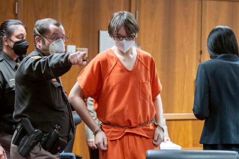 A teenage boy wearing glasses, a white mask over his face and dressed in an orange prison jumpsuit is led out of a court with his hands cuffed to a chain around his waist.