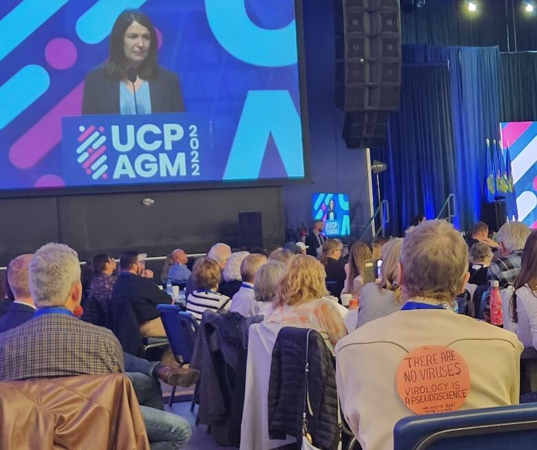 People sit watching a politician speak on stage, and a larger video version of her.