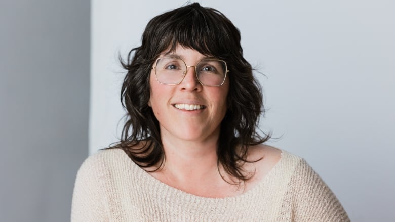 A woman sits in front of a grey background. She wears a beige sweater and circular glasses.