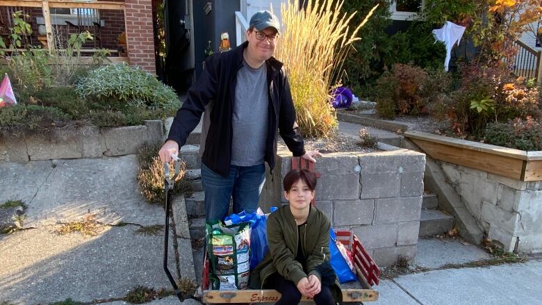 A man and child stand in front of a row of houses. They have a wagon filled with groceries.