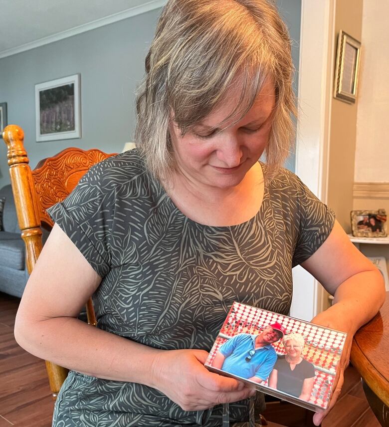 A woman sits on a chair, leaning on a table on the right-hand side of the photo. She looks down at a framed photo in her hands, which shows a man and a woman, both smiling.