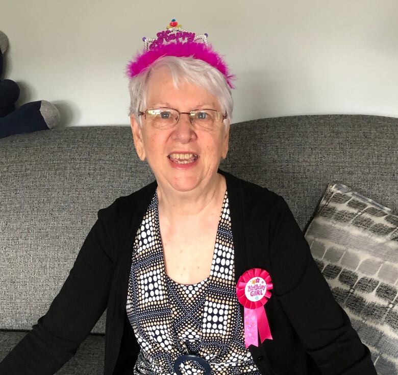 An older woman is smiling. She wears both a birthday headpiece and ribbon.