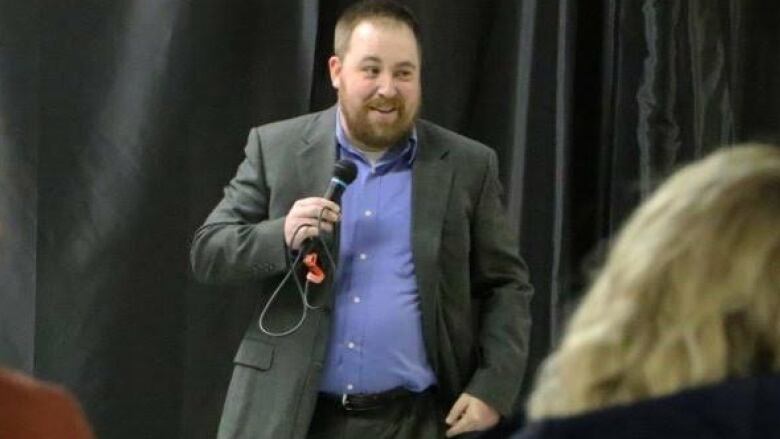 A man in a suit holds a microphone in front of an audience.