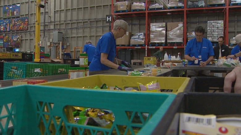 Volunteers wearing blue shirts and gloves sort through packaged food donations in a warehouse. 