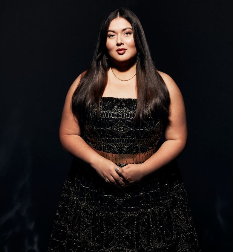 A woman wearing a black dress stands in front of a black background.