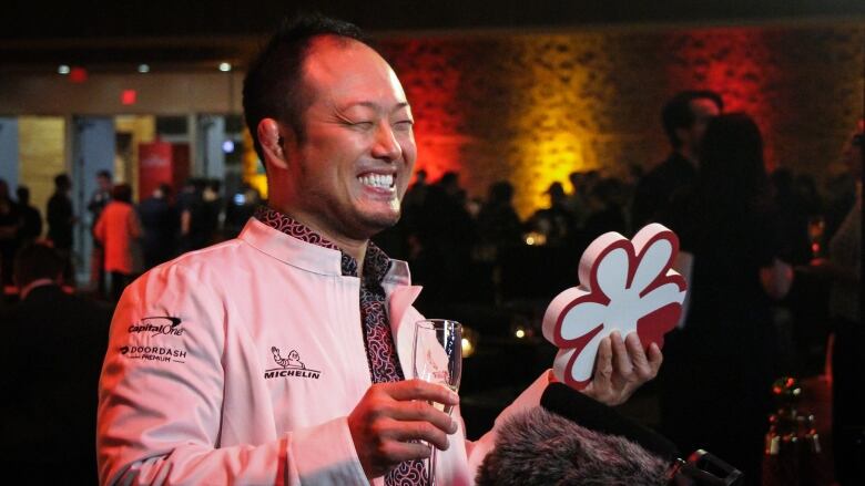 A smiling man in a white jacket holds a glass of champagne and a white star symbolizing his restaurant's entry to the Michelin Guide.