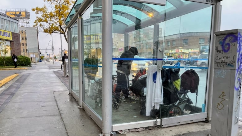 Several people gather in a bus shelter with glass walls on a busy city street.