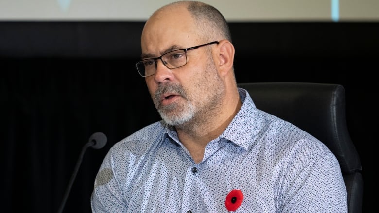 A man in a dress shirt and glasses speaks into a microphone.