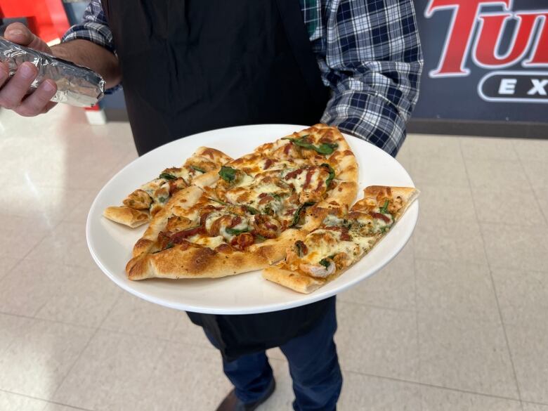 A man holds a plate covered in a flatbread topped with meat, cheese and vegetables.