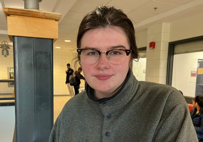 A brown-haired woman is wearing glasses and a grey sweater. She is leaning on a railing. 