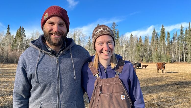 A man and woman wear toques in a cow field on a brisk fall day