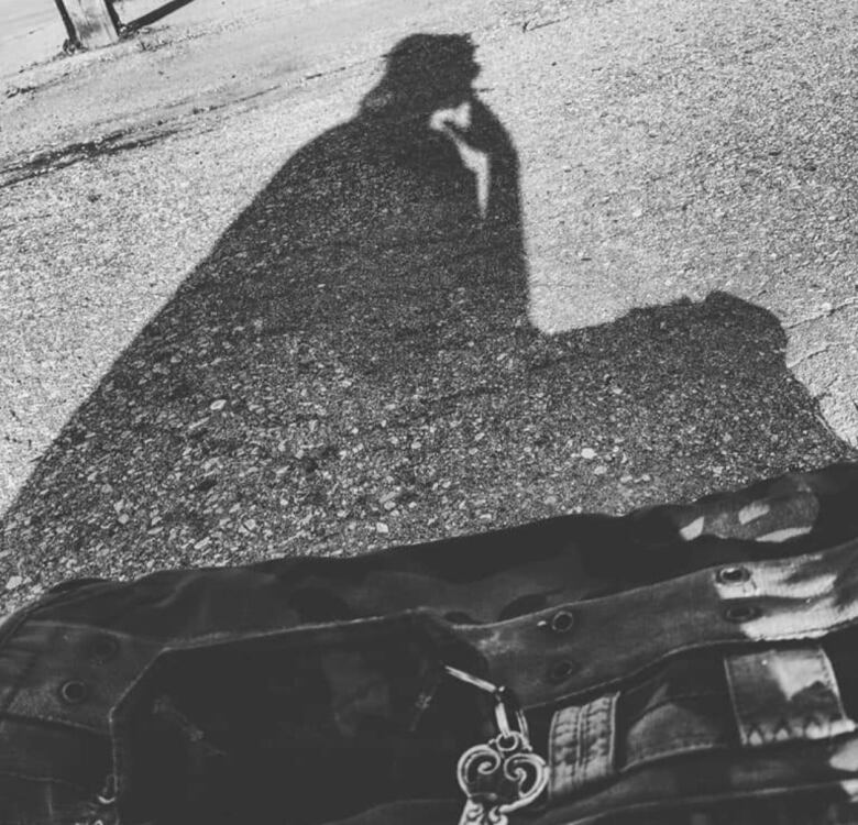 Shadow of a woman sitting sideways on pavement and smoking a cigarette, with a dark duffel bag in the foreground.