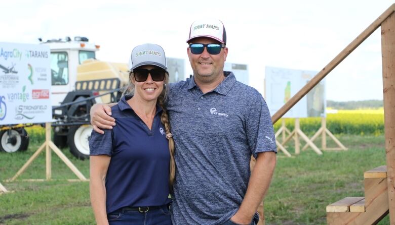 A man and woman wearing ballcaps that have branding for 'SWAT Maps' on them stand with their arms around one another. In the background, you can see a farmer's field and machinery.