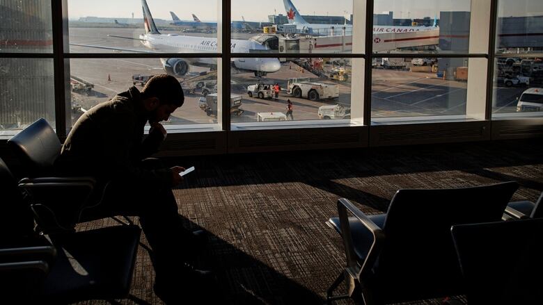 A man sits in the waiting area of the airport with a smartphone in his hand. He's backlit by a window that shows an airplane being readied for boarding. 