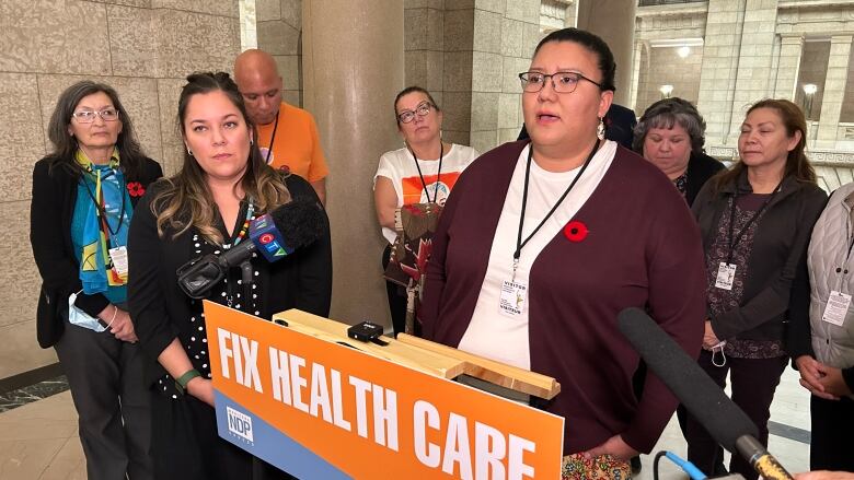 A woman speaks at a podium that says, 'fix health care' as a group of people surround her.