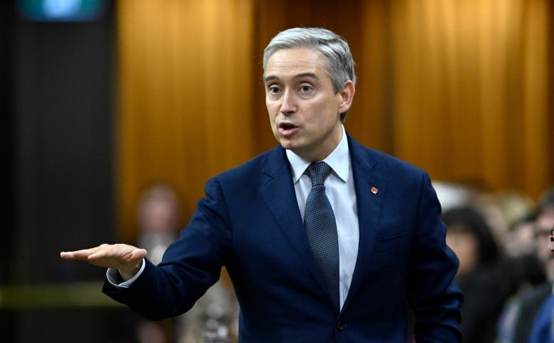 A man with white hair and a blue suit gestures with is right hand.