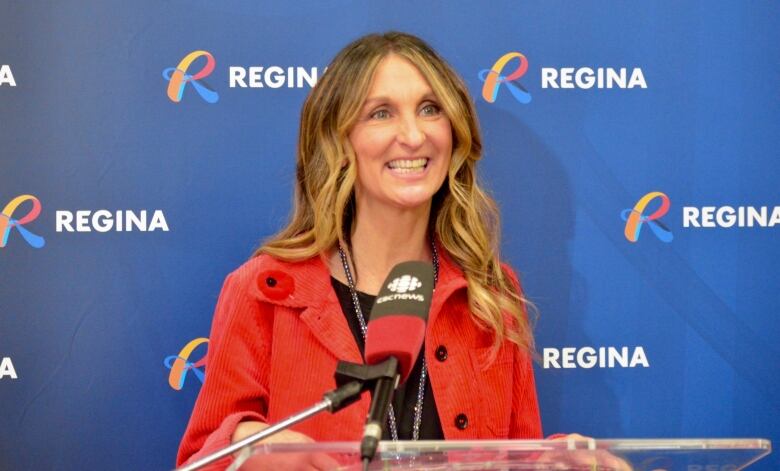 A smiling woman in a red blazer stands at a podium in front of a backdrop with the City of Regina logo.