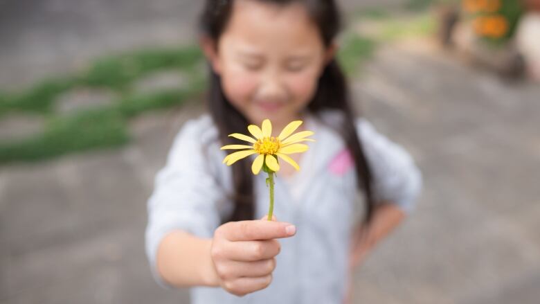 A blurred image of a smiling young girl with pigtails holding out a yellow flower in her hand.