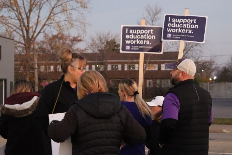 Five people with their backs to the camera are gathered. one is holding a sign that says 'I support education workers.' 