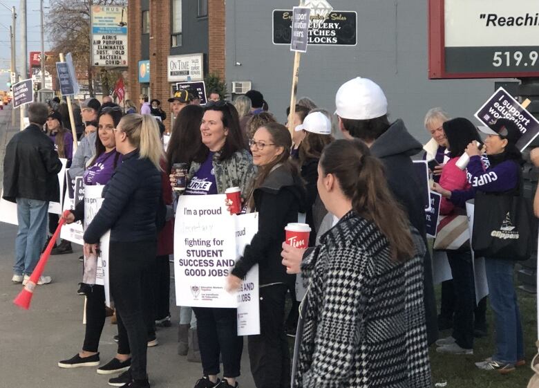 A group of people stand on a sidewalk holding signs and coffees. 