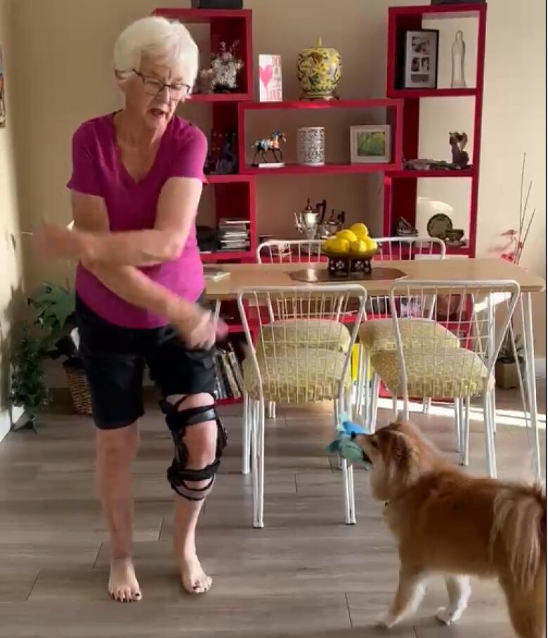 A woman wearing a knee brace dances in a dinning room while a small dog looks on.