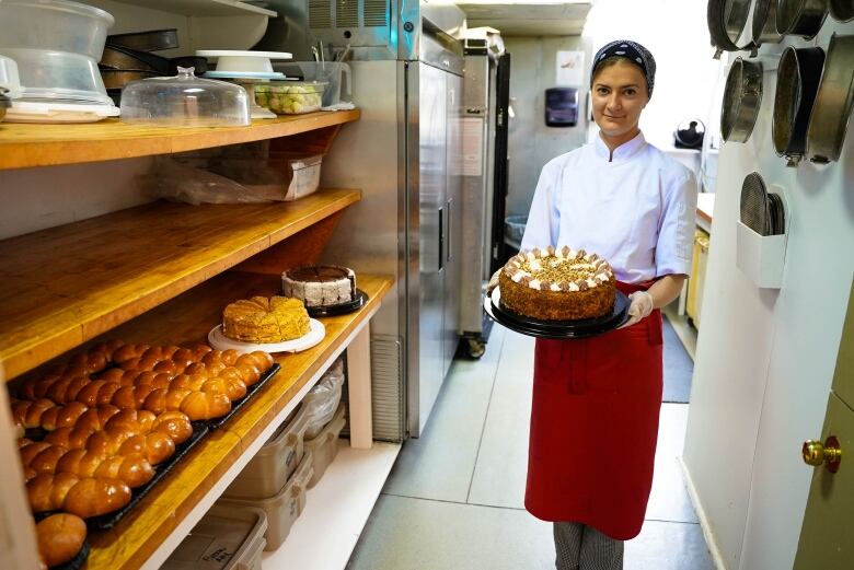 A woman stand sin a kitchen with racks of fresh bread, presenting a newly-decorated cake.