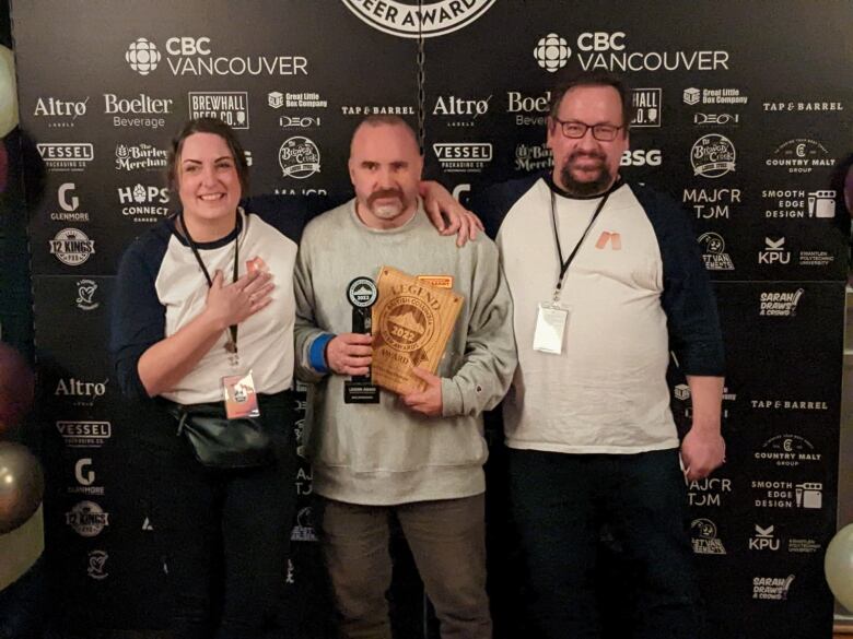 Three people pose in front of an awards ceremony backdrop. The person in the centre, a white man with close-cropped hair, is carrying trophies.